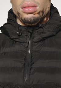 Giubbotto imbottito nero con zip frontale, cappuccio regolabile e texture lucida. Caratterizzato da cuciture orizzontali e dettagli di design minimal.