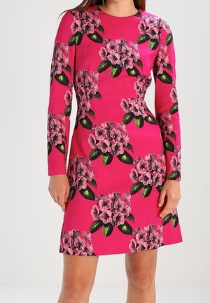 Frau trägt ein hellpinkes langärmeliges Kleid mit großem Blumenmuster in Pink- und Grüntönen, steht mit entspannten Händen.