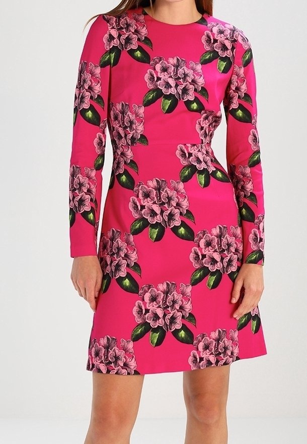 Femme portant une robe rose vif à manches longues avec un grand imprimé floral dans des nuances de rose et de feuilles vertes, debout, les mains détendues.