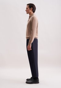 Mann, der seitlich steht, trägt ein beiges Langarm-Polo, marineblaue Hose, schwarze Socken und schwarze Leder-Schnürschuhe vor einem schlichten Hintergrund.
