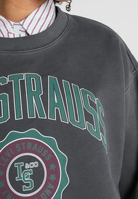 Sudadera gris oscuro con un logo impreso en verde y marrón que presenta el texto "LEVI STRAUSS" y un diseño de emblema redondo. Material de algodón.