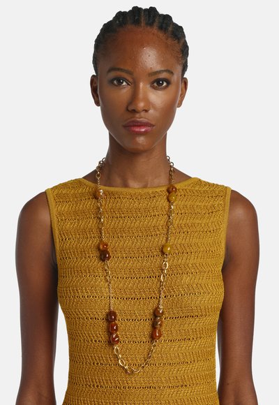 Fonott hajú nő, mustársárga, ujjatlan kötött felsőt visel, hosszú aranylánccal, borostyánszínű gyöngyökkel.