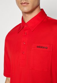 Polo roșu confecționat dintr-un material neted, având un guler clasic și o plachetă cu nasturi. Dispune de mâneci scurte și o buzunar cu patch, decorate cu un logo brodat în negru.