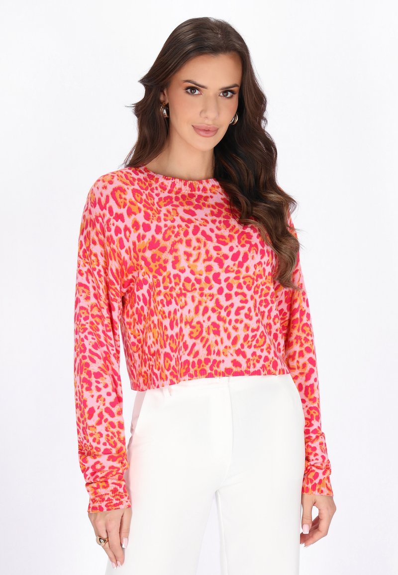 Frau mit langen braunen Haaren, die Creolen trägt, ein langärmliges Oberteil mit rosa-orangefarbenem Leopardenmuster und weiße, hoch taillierte Hosen.