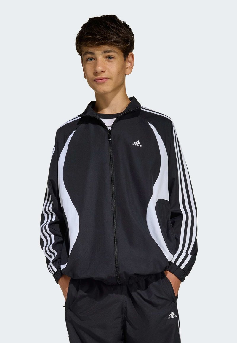 Dospievajúci chlapec oblečený v čierno-bielom teplákovom súprave Adidas s rukami v vreckách, stojaci pred jednoduchým svetlým pozadím.