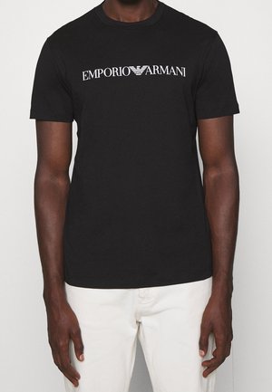 Homme portant un t-shirt noir à manches courtes avec le logo blanc "EMPORIO ARMANI" et un pantalon blanc, debout devant un fond uni.