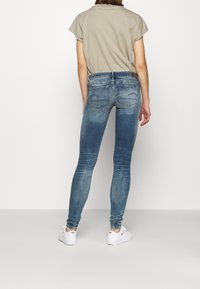 Persona vistiendo jeans skinny azules desgastados, camiseta beige y zapatillas Nike blancas, de pie de espaldas contra un fondo blanco.