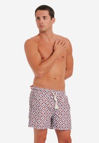 Bruna mönstrade badshorts med blå diamantformer, med en dragsko i midjan. Modellen poserar med en hand på axeln.