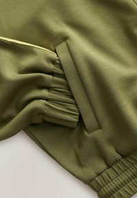 Manche de vêtement vert olive avec poignet élastique, poche latérale et détail de passepoil jaune près de la couture de l'épaule.