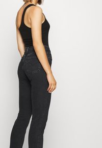 Mujer con una blusa sin mangas negra y jeans de tiro alto gris oscuro de pie contra un fondo blanco liso.
