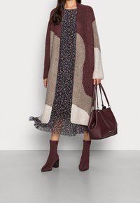 Pletený oversized cardigan v burgundské a béžové barvě, držený přes tmavé květinové šaty. Burgundské podpatkové boty a odpovídající kabelka dotvářejí vzhled.