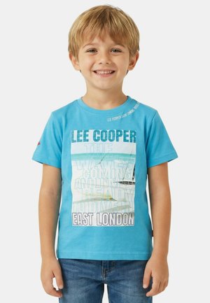 Jeune garçon souriant aux cheveux blonds portant un t-shirt bleu Lee Cooper avec un voilier et le texte "East London".