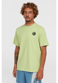 Camiseta de algodón verde claro con cuello redondo clásico y mangas cortas. Presenta un logo circular negro en el lado izquierdo del pecho.