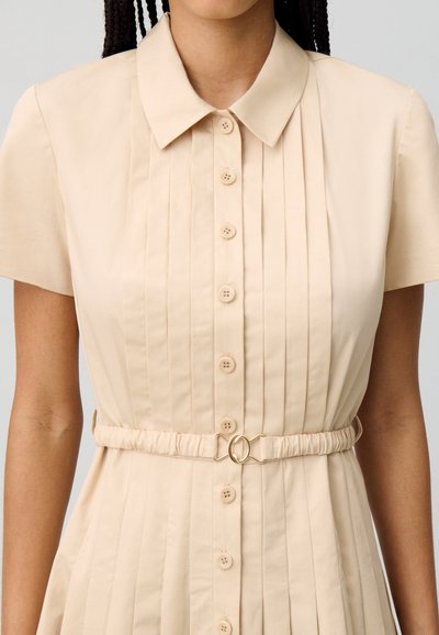 Vestido beige con botones, de mangas cortas, diseño plisado y cinturilla elástica con un detalle de hebilla ovalada en tono dorado.