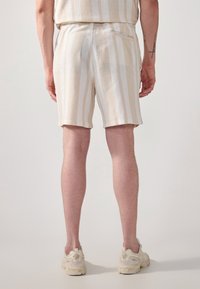 Pantalones cortos de rayas beige con un tejido texturizado, que cuentan con una cintura elástica y un bolsillo trasero. El modelo lleva zapatillas de color claro.