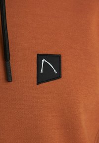 Látka barvy rezavě hnědé s černou záplatou nesoucí bílý geometrický vzor a černým stahovacím provázkem s nápisem "CHASIN." na konci.