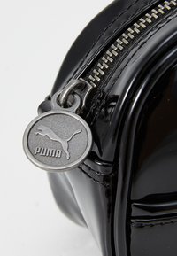 Svart lackläderväska med en silverdragkedja och en rund silver Puma-logotyp som dragkedjering. Slät textur med sydda accenter.
