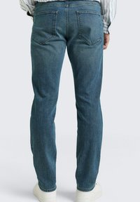 Man draagt blauwe jeans met rechte pijpen en witte sneakers, staat met handen langs zijn zij op een effen achtergrond.