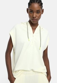 Kobieta z warkoczami, ubrana w bezrękawnik w kolorze kremowym z zamkiem błyskawicznym i sznurkiem, trzyma ręce w kieszeniach pasujących kremowych spodni.