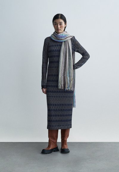 Gebreide jurk met een geometrisch patroon in donkerblauw en grijs. Gecombineerd met een multicolor zigzag sjaal en bruine kniehoge laarzen.