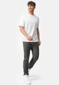Camiseta blanca de manga corta combinada con pantalones ajustados gris y zapatillas blancas. El modelo está en una pose casual contra un fondo claro.
