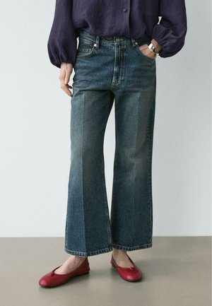 Persona che indossa jeans a gamba larga blu scuro, una camicetta a maniche lunghe viola scuro, ballerine rosse e un orologio da polso argentato.