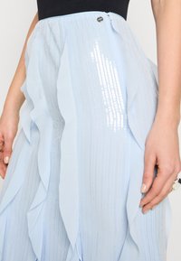 Jupe superposée bleu clair en tissu transparent avec des rayures en sequins, présentant des bords froncés et une fermeture éclair sur le côté.