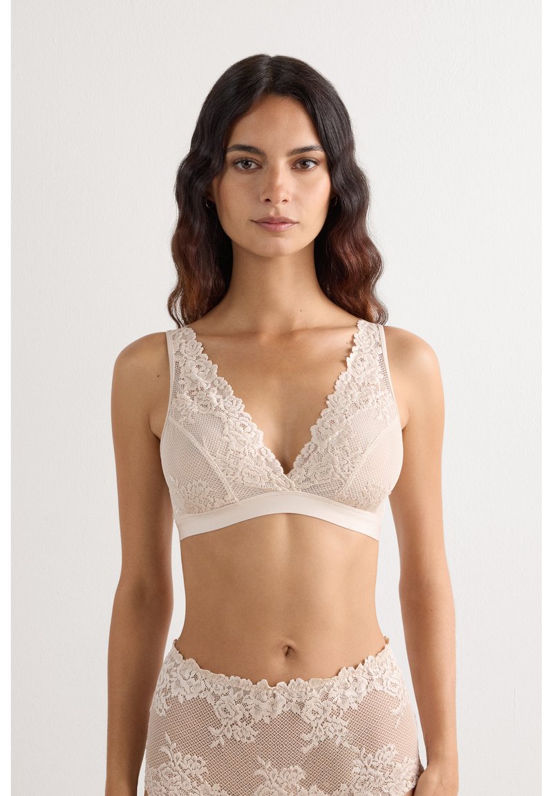Kvinde med langt mørkt hår iført matchende beige blonde-bh og højtaljede blonde-trusser, der vender fronten mod en ensfarvet lys baggrund.