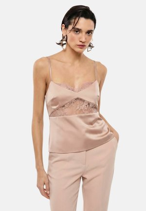 Donna che indossa una canottiera in raso color cipria con pannello in pizzo e pantaloni coordinati, posando con una mano in tasca davanti a uno sfondo bianco.