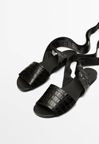 Schwarze flache Sandalen mit einem strukturierten Krokodilmuster und einem breiten Riemen, ausgestattet mit einer glatten Sohle und langen, gekreuzten Stoffbändern.