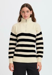 Maglione a maglia a righe crema e nere. Collo alto con chiusura a zip, polsini e orlo a costine. Tessuto morbido, vestibilità ampia.