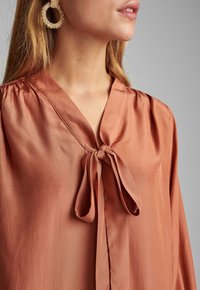 Frau, die eine rostfarbene Satinbluse mit einer gebundenen Schleife am Kragen und texturierten goldenen Creolen trägt, vor einem neutralen Hintergrund.