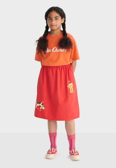 Niña con cabello trenzado que lleva una camiseta naranja de "Bobo Choses", falda roja con diseños de granjero y vaca, calcetas a rayas y zapatillas rojas.