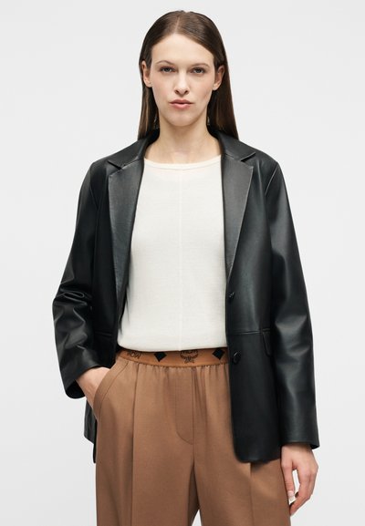 Femme portant un blazer en cuir noir sur un haut blanc et un pantalon taille haute marron avec une ceinture élastique, debout avec une main dans la poche.
