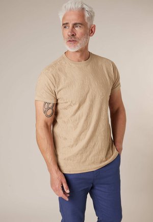 Man met wit haar en baard, gekleed in een beige T-shirt met patroon en een blauwe broek, met een tatoeage zichtbaar op zijn linker bovenarm.