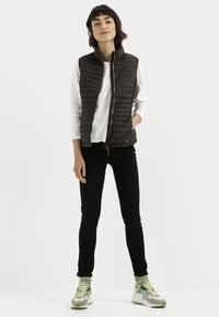 Zwarte gewatteerde vest met hoge kraag, ritssluiting, gedragen over een witte longsleeve shirt. Gecombineerd met zwarte jeans en meerkleurige sneakers.