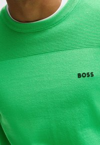 Grön stickad tröja med en texturerad horisontell randdesign vid bröstet och varumärket "BOSS" i svart.