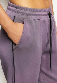 Lila Jogginghosen aus einem weichen Stoff, mit elastischem Bund, seitlichen Taschen und schwarzen Kordelzug-Akzenten. Insgesamt glatte Textur.