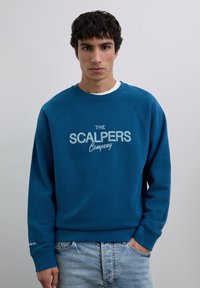 Jeune homme portant un sweat-shirt bleu avec le texte "The Scalpers Company", un jean bleu clair, se tenant devant un fond gris clair uni.