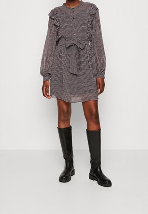 Langärmliges Kleid mit Rüsche an den Schultern, Knopfleiste, tailliertem Gürtel und Pünktchenmuster in Marineblau und Weiß; kombiniert mit knielangen schwarzen Stiefeln.