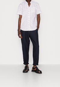 Chemise blanche à manches courtes avec des pois rouges, associée à un pantalon en lin bleu marine et des sandales noires à lanières. Coupe décontractée et poignets de pantalon retroussés.