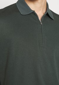 T-shirt polo vert foncé avec un col zippé et un col texturé, mettant en valeur une finition en tissu lisse et une coupe ajustée.