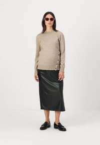 Beige neulepusero, jossa on nappiyksityiskohtia sivussa, yhdistettynä kiiltävään mustaan nahkaiseen midi-hameeseen ja mustiin loafer-kengiksiin. Vaaleat aurinkolasit.