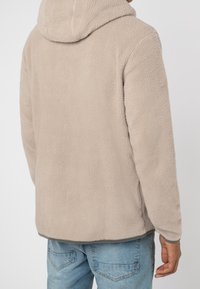 Homme portant un sweat à capuche en polaire beige et un jean bleu clair, vu de dos sur un fond uni.