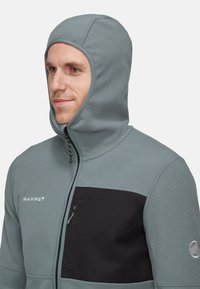 Grau Fleece-Jacke mit einer strukturierten Kapuze, schwarzem Brustpanel und Reißverschlüssen. Verfügt über ein Logo an der Seite und eine Tasche mit einem Zugband.