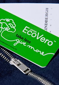 Vihreä EcoVero-tunnus valkoisella tekstillä, jossa on puugrafiikka, tumman kankaan päällä, jossa on hopeinen vetoketjuyksityiskohta ja valkoinen kortti taustalla.