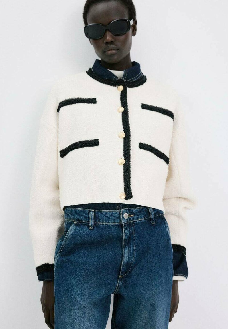 Personne portant des lunettes de soleil noires, un cardigan blanc court avec bordure noire et boutons dorés, superposé à une chemise en denim foncé et un jean bleu taille haute.