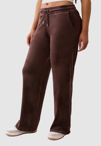 Osoba w brązowych, szerokich spodniach z weluru z elastycznym pasem i bocznymi kieszeniami, w połączeniu z białymi i beżowymi trampkami.