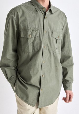 Homme portant une chemise vert olive à manches longues boutonnée avec deux poches poitrine et un pantalon beige.