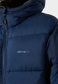 Chaqueta acolchada azul marino con cierre de cremallera y capucha. Presenta un diseño acolchado y un logo metálico de Carhartt en el lado izquierdo del pecho.